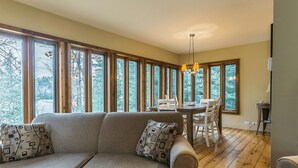 A cozy living room with a sofa and a dining table, large windows with a view of trees, and wooden flooring.