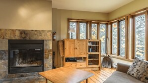 A cozy living room with a stone fireplace, wooden furniture, and large windows offering a view of autumn foliage.