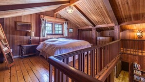 Un dormitorio de cabaña de madera con cama, mesita de noche, ventana con cortinas, escalera y estantería.