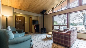 A cozy living room with a fireplace, a television, and large windows offering a view of the outdoors. - Welcome to the Chalets du Lac Grenier (CHERTSEY)