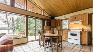 A spacious kitchen with wooden cabinets, a dining table with chairs, and a view of the outdoors through large windows. - Welcome to the Chalets du Lac Grenier (CHERTSEY)