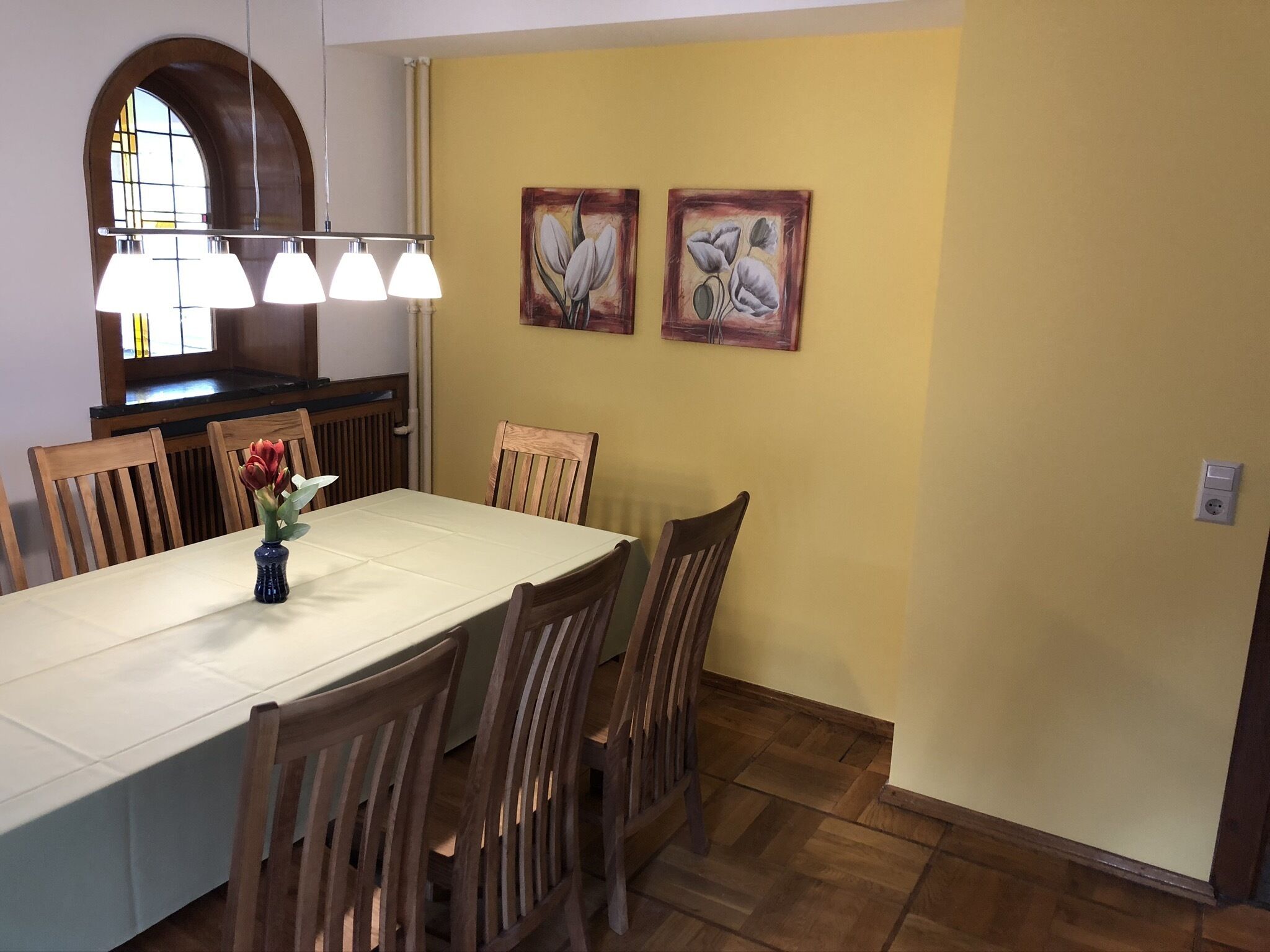 A dining area with a long table, wooden chairs, and two paintings on the wall.