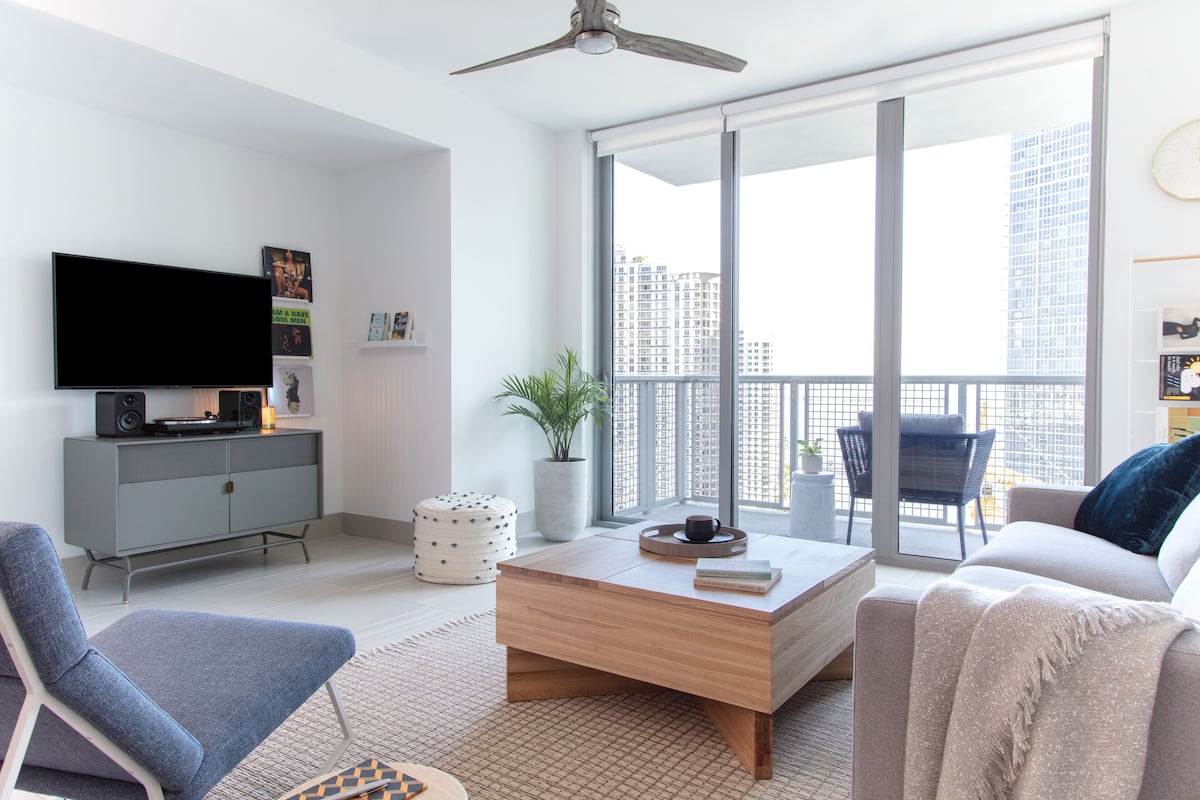 A modern living room with a flat-screen TV, a grey cabinet, a wooden coffee table, a blue armchair, a beige sofa, and a large window with a city view.