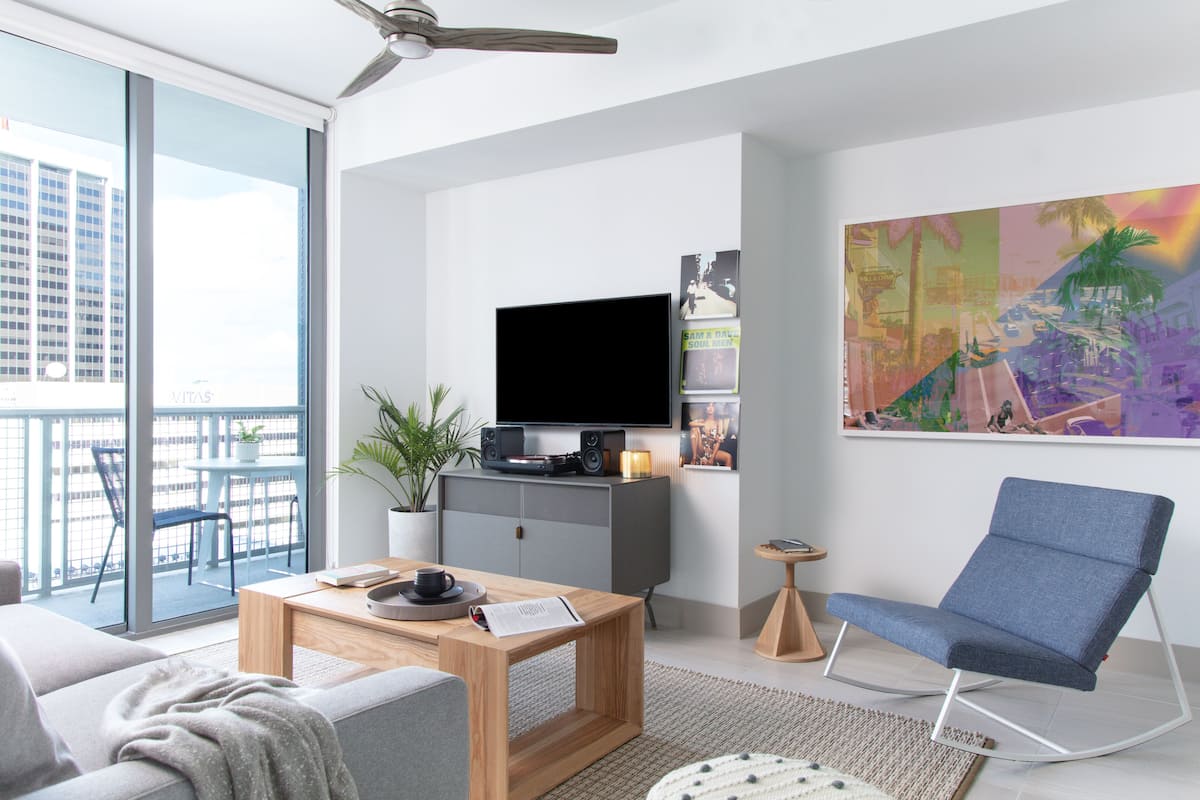 A modern living room with a flat-screen TV, a blue chair, a wooden coffee table, and a large window overlooking a cityscape.
