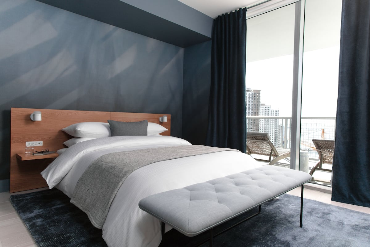 A modern bedroom with a large bed, a wooden headboard, a bedside table, and a view of the city through a large window.