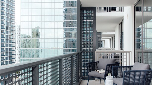 A balcony with wicker furniture, a white round table, and a view of tall buildings. - Mint House Miami - Downtown (Miami)