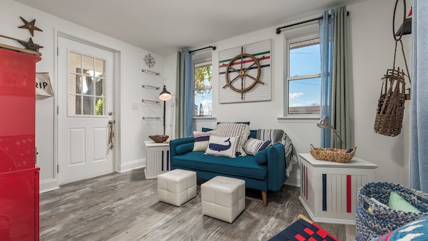 A living room with a teal sofa, white ottomans, a red cabinet, and nautical decor including a ship's wheel and a star. - 303 Massachusetts - Adorable one bedroom cottage close to the beach! (Lewes)