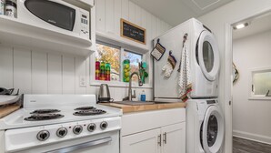 A kitchen with a stove, microwave, and washing machine. There is a window with a view of trees and a chalkboard sign above the window. - 303 Massachusetts - Adorable one bedroom cottage close to the beach! (Lewes)