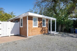 A small wooden structure with a white roof and a covered porch, surrounded by gravel and trees. - 303 Massachusetts - Adorable one bedroom cottage close to the beach! (Lewes)