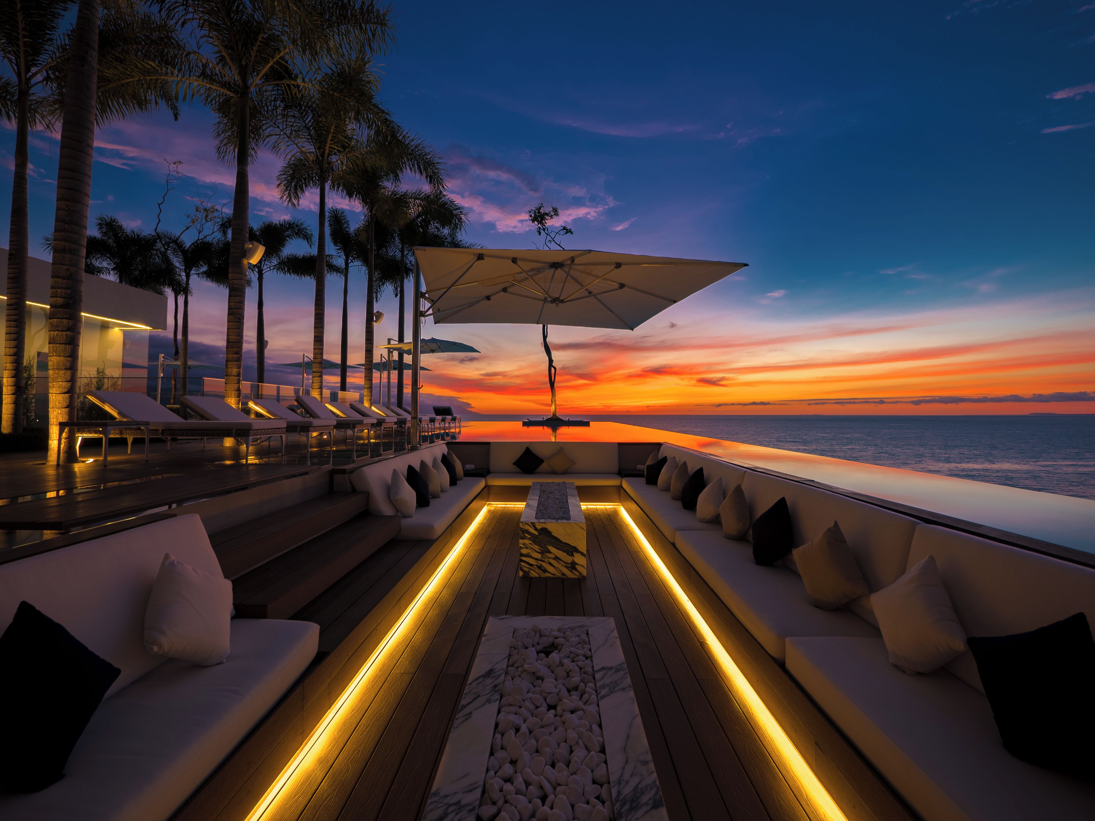 A sunset view from a deck with lounge chairs and a pool.
