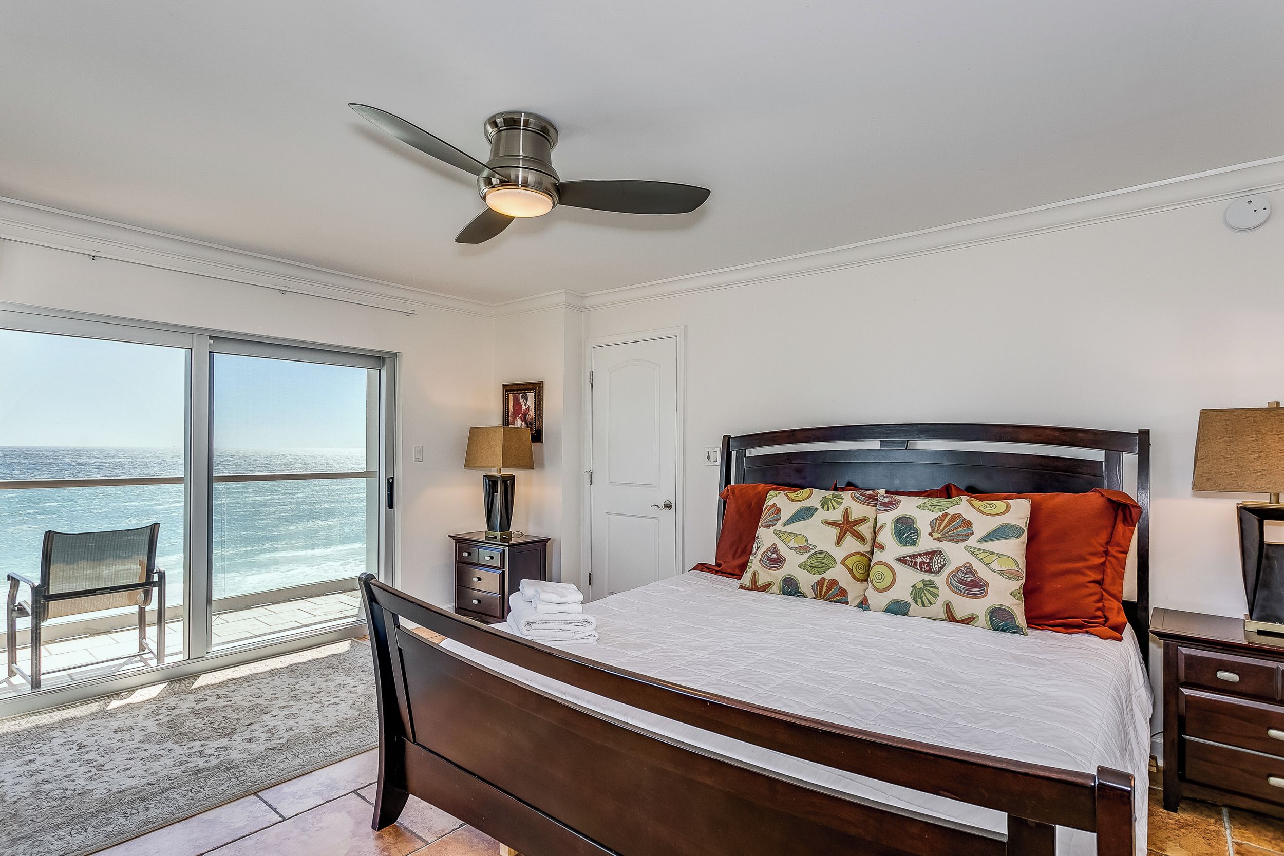 A bedroom with a bed, a ceiling fan, a nightstand, a lamp, a chair, and a view of the ocean.