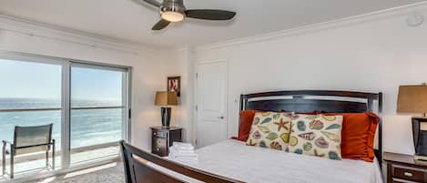 A bedroom with a bed, a ceiling fan, a nightstand, a lamp, a chair, and a view of the ocean.