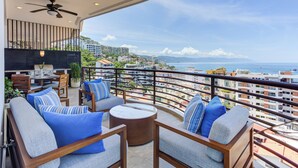 A balcony with outdoor seating, a ceiling fan, and a view of the city and sea.
