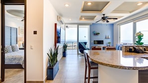 A modern kitchen with a central island, a dining area with a blue chair, and a view of the ocean through large windows.