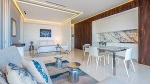 Une chambre d’hôtel moderne comprenant un lit, un coin salon avec un canapé et une table basse, une table à manger avec des chaises, et un coin cuisine avec un pare-feu en marbre.