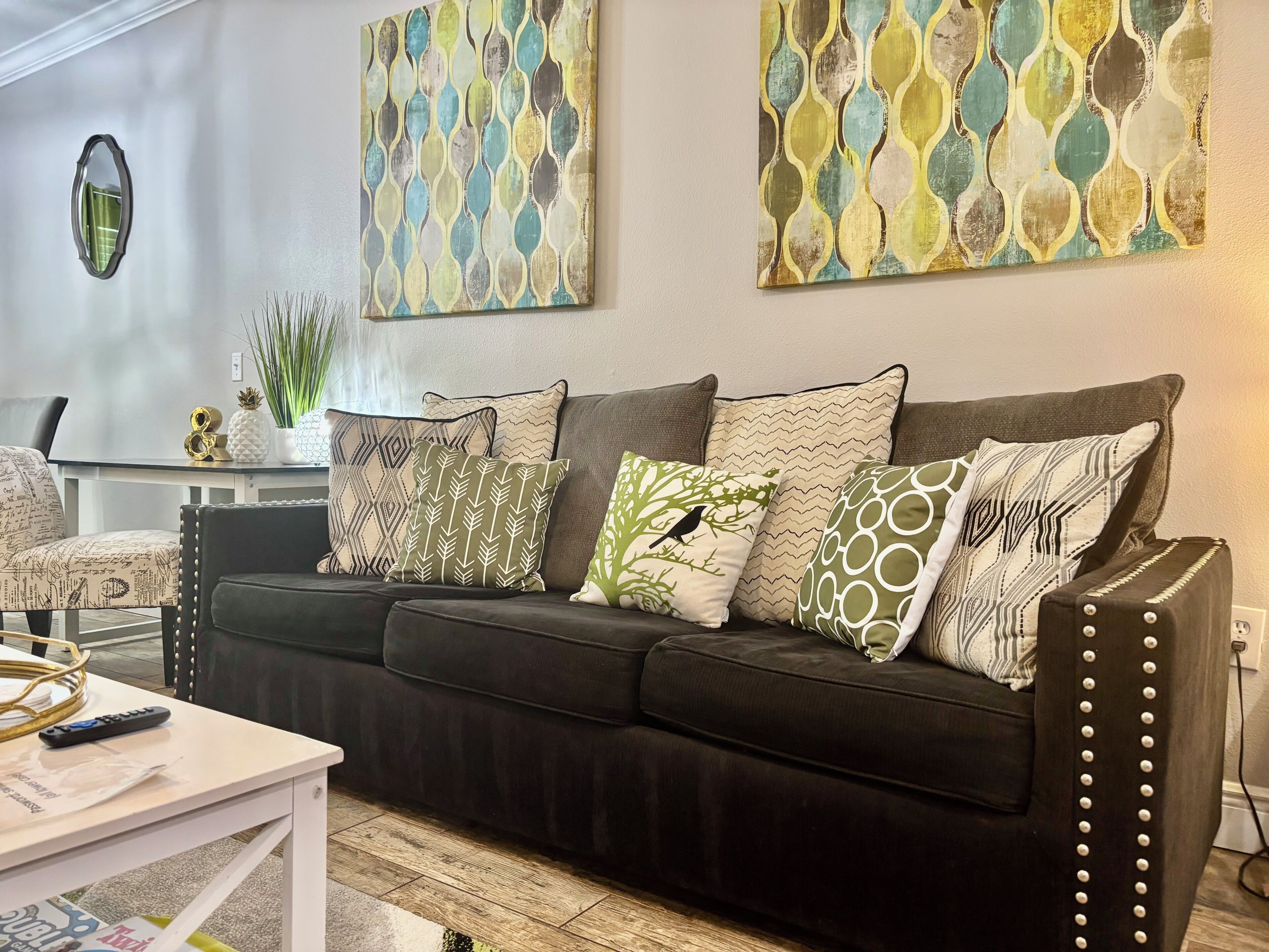 A living room with a dark-colored sofa adorned with various patterned cushions, a white coffee table, and a wall-mounted mirror.