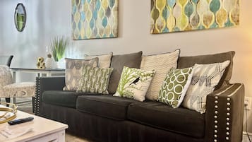 A living room with a dark-colored sofa adorned with various patterned cushions, a white coffee table, and a wall-mounted mirror.