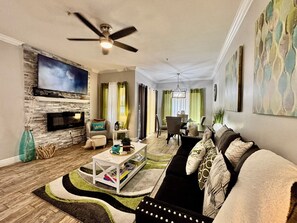 A living room with a fireplace, a ceiling fan, a television, and a dining area with a chandelier. - SHEEK & GLAM VACATION BEACH CONDO! HEATED POOL,GYM, TENNIS, 1st FLOOR CONDO (Seminole)