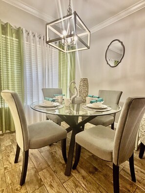 A dining area with a round glass table, four chairs, a chandelier, and a mirror on the wall.