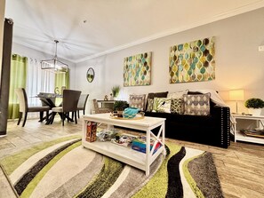 A living room with a sofa, a coffee table, a dining area with chairs, and artwork on the walls.