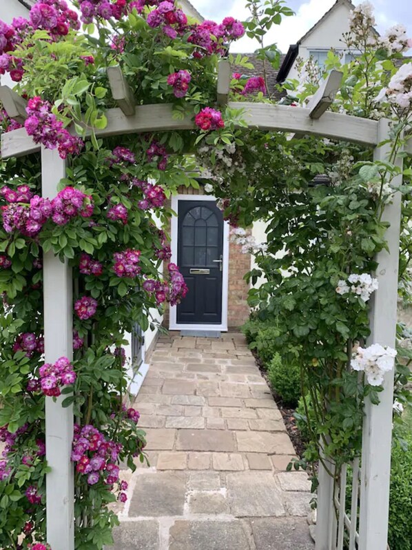 A stone pathway leading to a black door, surrounded by a wooden archway with pink flowers. - Cotswolds Blenheim Burford Oxford Bibury Minster Mill Diddly Farmers Dog Soho  (Minster Lovell)