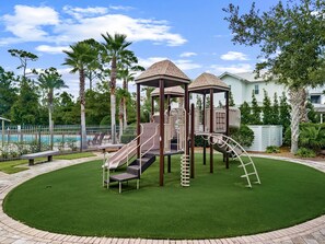 Ein Spielplatz mit Rutsche und Klettergerüsten, umgeben von einer gepflegten Rasenfläche und einem Pool im Hintergrund.