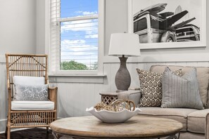 A room with a wicker chair, a round coffee table with a bowl, a sofa with patterned cushions, and a lamp.