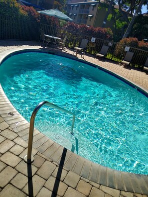 A clear swimming pool with a metal ladder, surrounded by a paved deck and a fence.