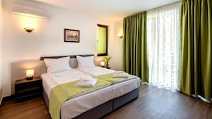 Habitación de hotel con una cama grande, dos mesitas de noche con lámparas, un cuadro en la pared y una ventana con cortinas verdes.