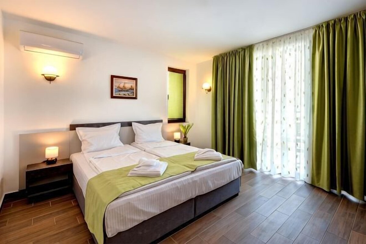 A hotel room with a double bed, bedside lamps, a nightstand, a green curtain, and a window with curtains.