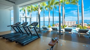Une salle de sport équipée de tapis de course et d’un banc, avec vue sur une piscine bordée de chaises longues et de palmiers.