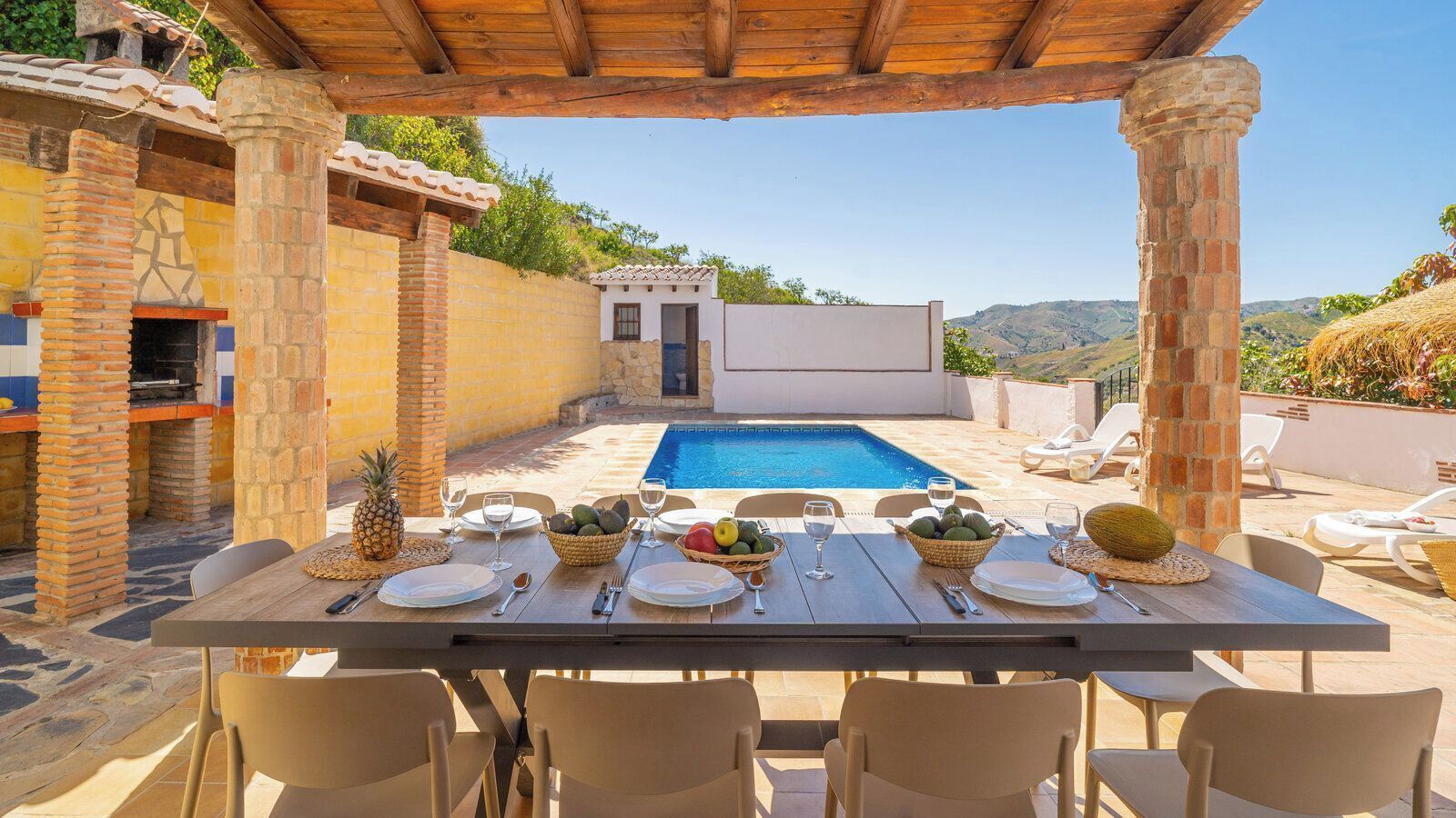 An outdoor dining area with a pool, stone pillars, and a view of the hills.