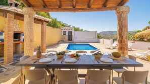 Un área de comedor al aire libre con piscina, pilares de piedra y vistas a las colinas.