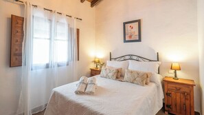 Una cama bien hecha con sábanas blancas, dos mesitas de noche con lámparas, un cabecero de madera y una ventana con cortinas de gasa.