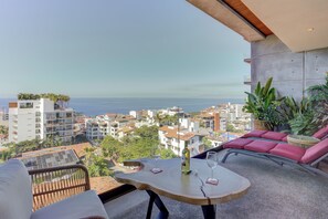 A balcony with a table, two chairs, and a bottle of wine. The view includes a coastal cityscape with buildings and palm trees.
