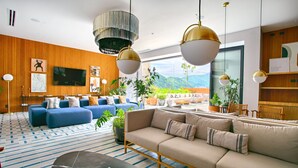 En moderne stue med en stor sofa, et sofabord og et fjernsyn. Der er krukker med planter, og der er udsigt til bjerge gennem vinduerne.