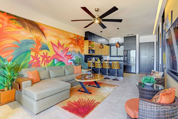 A modern living room with a sofa, coffee table, and chairs, a kitchen area, and a colorful mural on the wall.