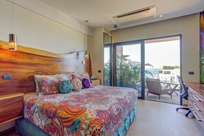 A bedroom with a large bed, a wooden headboard, a view of the outdoors through a sliding door, and a balcony with a chair and table.