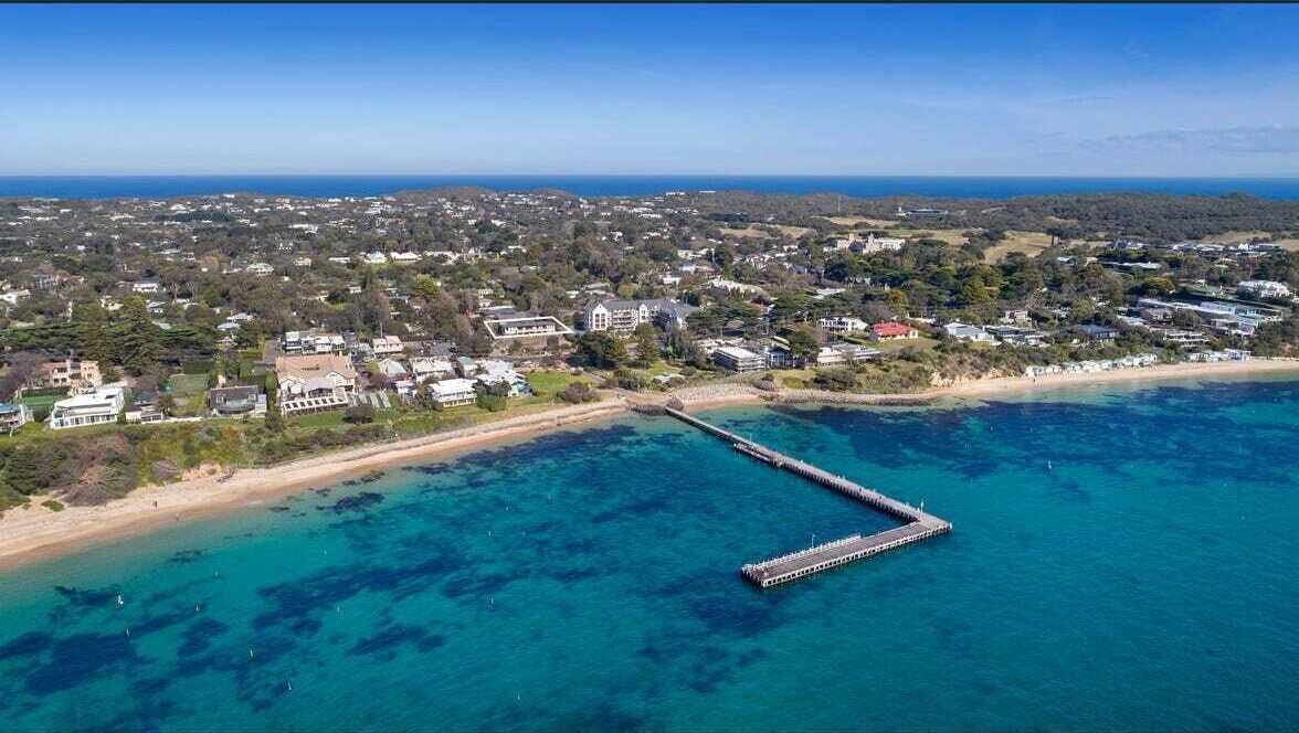 An aerial view of a coastal area with a pier extending into the sea, surrounded by residential buildings and a beach.