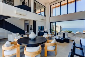 A modern living room with a round black table, white sofas, and wooden chairs. There is a large window with a view of the ocean and buildings. - Sail View Ocean Views, Rooftop Pool, Romantic Zone (Puerto Vallarta)
