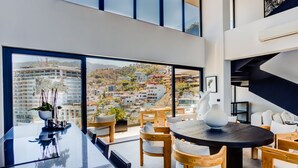 A modern dining area with a large window offering a city view, wooden furniture, and a black dining table. - Sail View Ocean Views, Rooftop Pool, Romantic Zone (Puerto Vallarta)