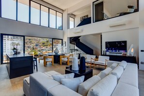 A spacious living area with a large sofa, a dining table with chairs, a kitchen area, and a staircase leading to an upper level. - Sail View Ocean Views, Rooftop Pool, Romantic Zone (Puerto Vallarta)