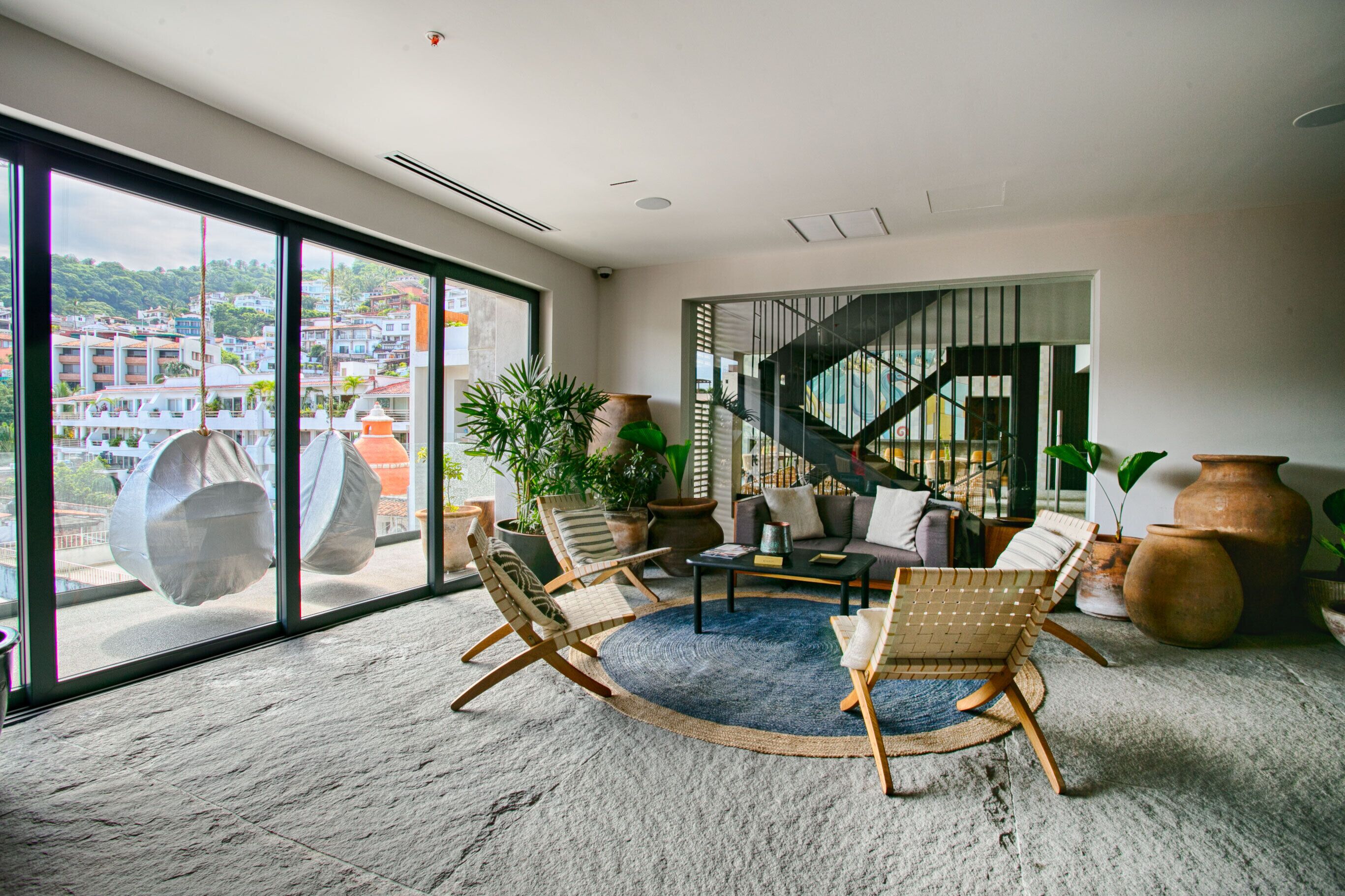 A spacious living room with large windows, a round rug, and a staircase leading to an upper floor.