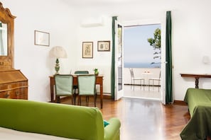 A room with a wooden dresser, a green sofa, a dining table with chairs, and a view of the sea through an open door.
