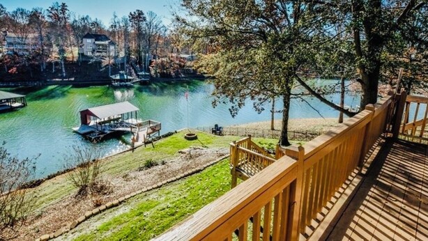 Una propiedad junto al lago con muelle de madera que da vista a un lago transparente, rodeada de árboles y un edificio distante.