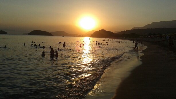 Ein Strandbild mit untergehender Sonne über dem Wasser, Menschen schwimmen und spazieren am Ufer entlang.