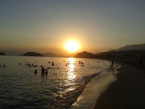 Uma praia com o sol se pondo sobre a água, pessoas nadando e caminhando pela orla.