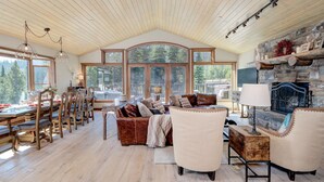 A spacious living room with a high ceiling, large windows offering a view of the forest, a fireplace with stone walls, and a dining area with wooden furniture. - Grey Moose Lodge | Ski-in/Out & Hot Tub (Big Sky)