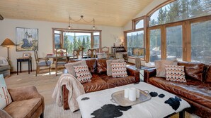 A spacious living room with a leather sofa, a dining area with a wooden table, and large windows offering a view of the forest. - Grey Moose Lodge | Ski-in/Out & Hot Tub (Big Sky)