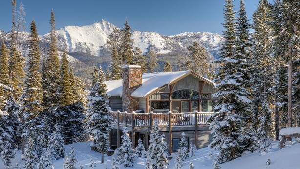 A lodge with a stone chimney, wooden deck, and a view of snow-covered mountains. - Grey Moose Lodge | Ski-in/Out & Hot Tub (Big Sky)